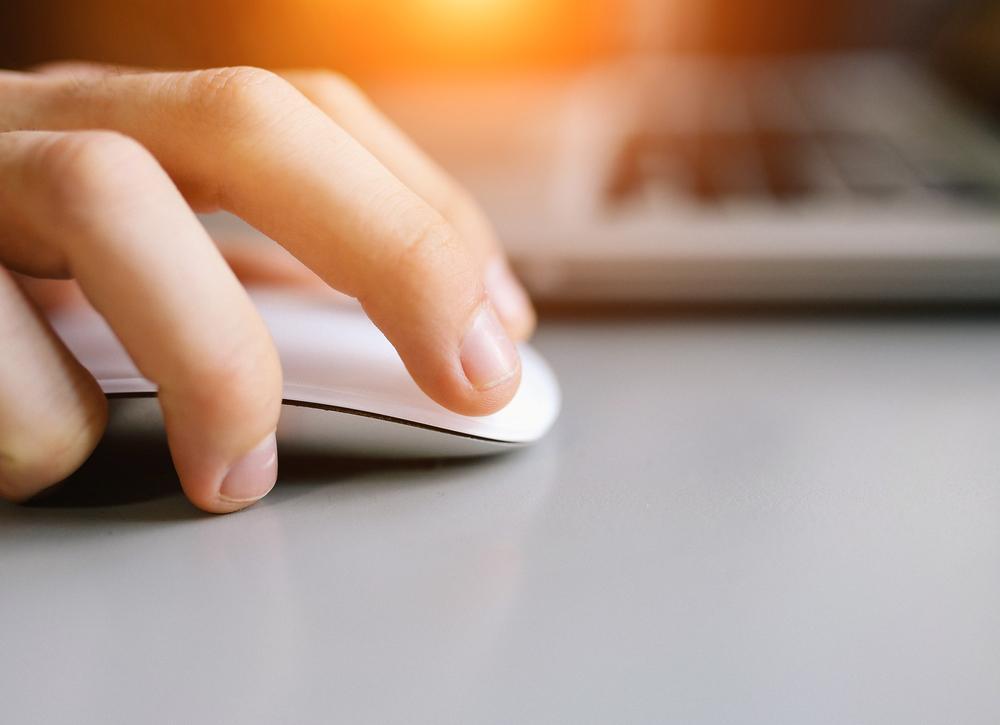 A close-up of a hand using a white computer mouse on a gray surface, with part of a laptop keyboard visible in the background and warm light shining from the top.