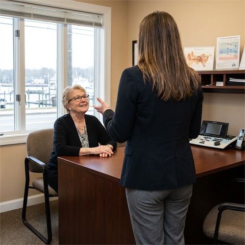 Two women are talking in an office. One, seated behind a desk, is smiling, while the other stands facing her. The office has a window, shelves with charts, and a phone on the desk.